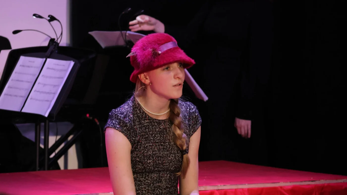 A Barney student in a pink hat sits and sings on stage.