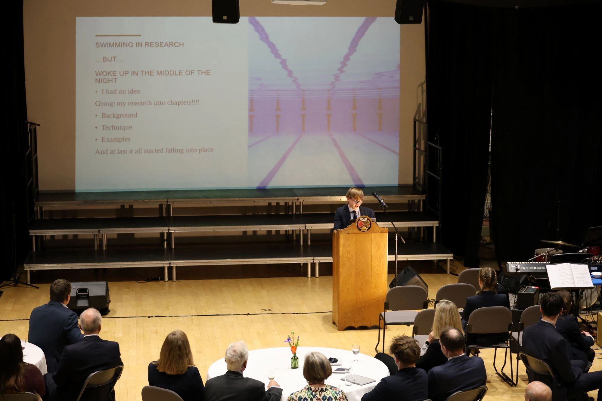 A student stands at a wooden podium giving a presentation titled 'Swimming in Research' to an audience seated at tables in a large hall.