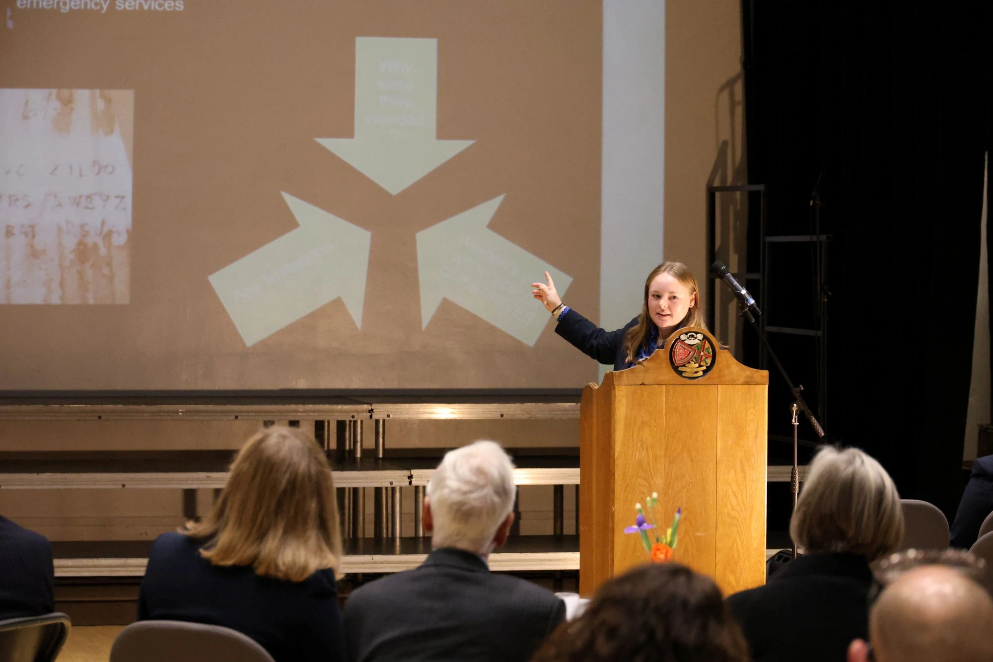  A student stands at a wooden podium giving a presentation, pointing toward a large projection screen displaying a diagram of three arrows pointing inward. 